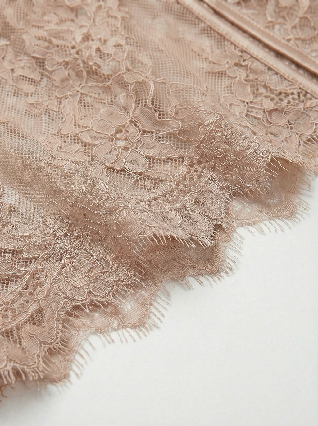 Detail shot of beige lace lingerie featuring a delicate eyelash lace hem and smooth satin piping. Sheer nude floral fabric highlighting elegant craftsmanship in women's intimates, bodysuits, and corset designs.