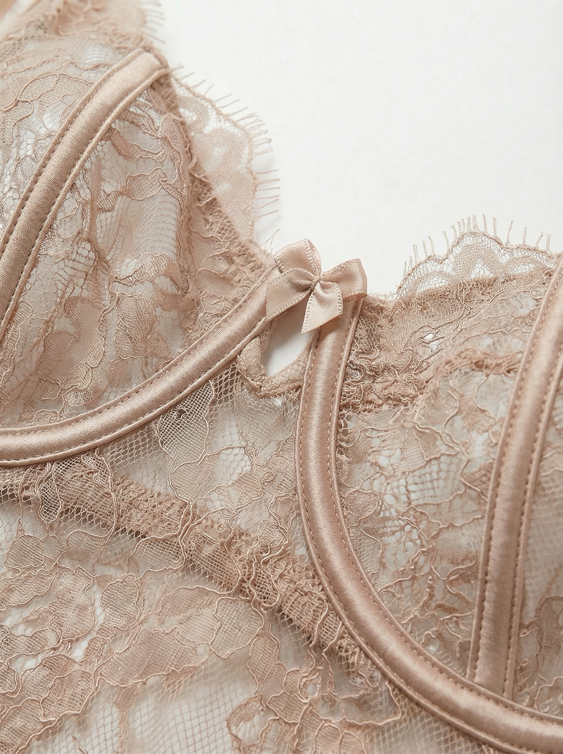 Close-up of a nude lace underwire bra cup and neckline. Features sheer beige floral lace, luxurious satin underwire casing, delicate eyelash lace trim, and a dainty satin center bow for elegant women's lingerie.