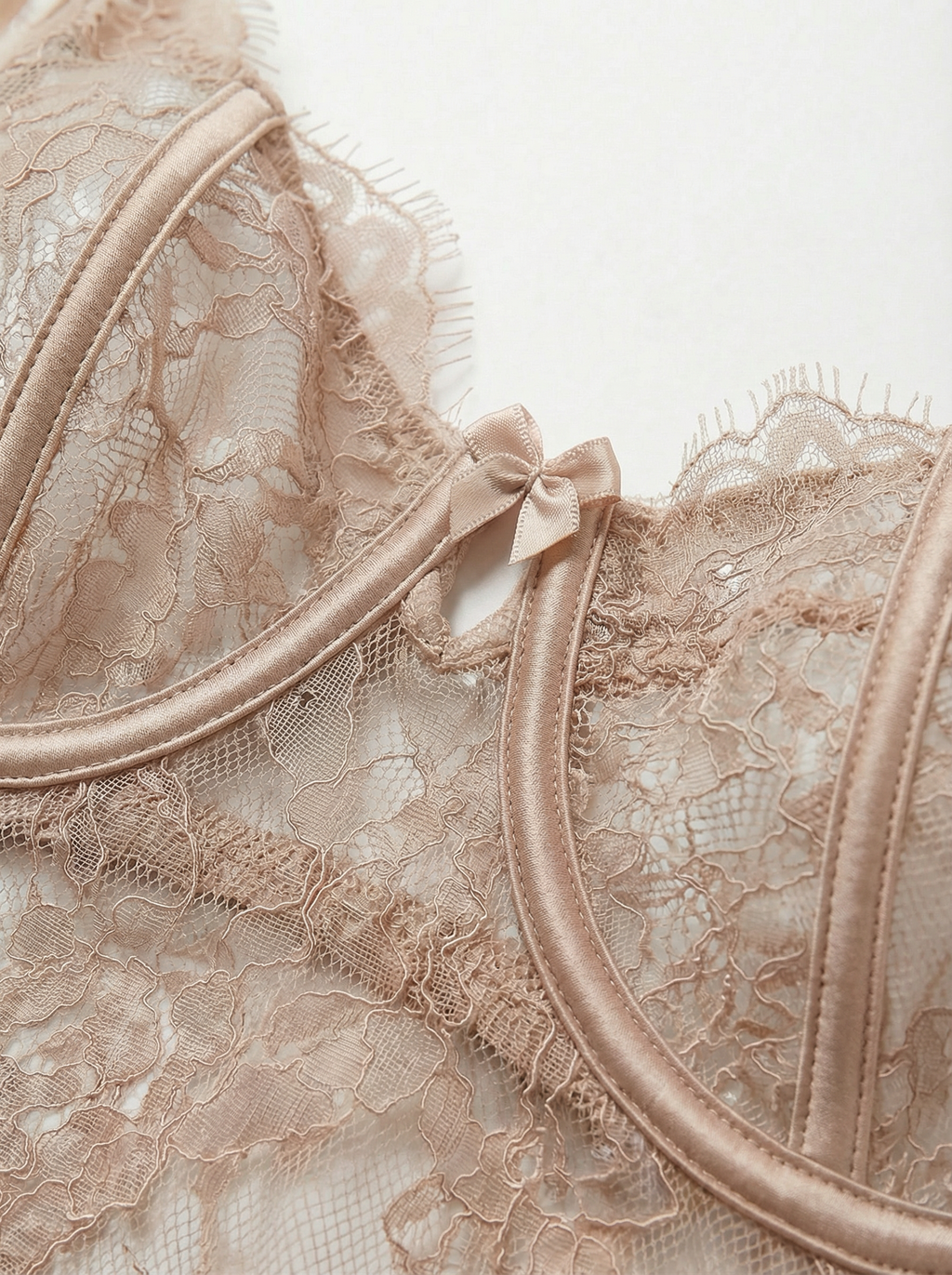 Close-up of a nude lace underwire bra cup and neckline. Features sheer beige floral lace, luxurious satin underwire casing, delicate eyelash lace trim, and a dainty satin center bow for elegant women's lingerie.