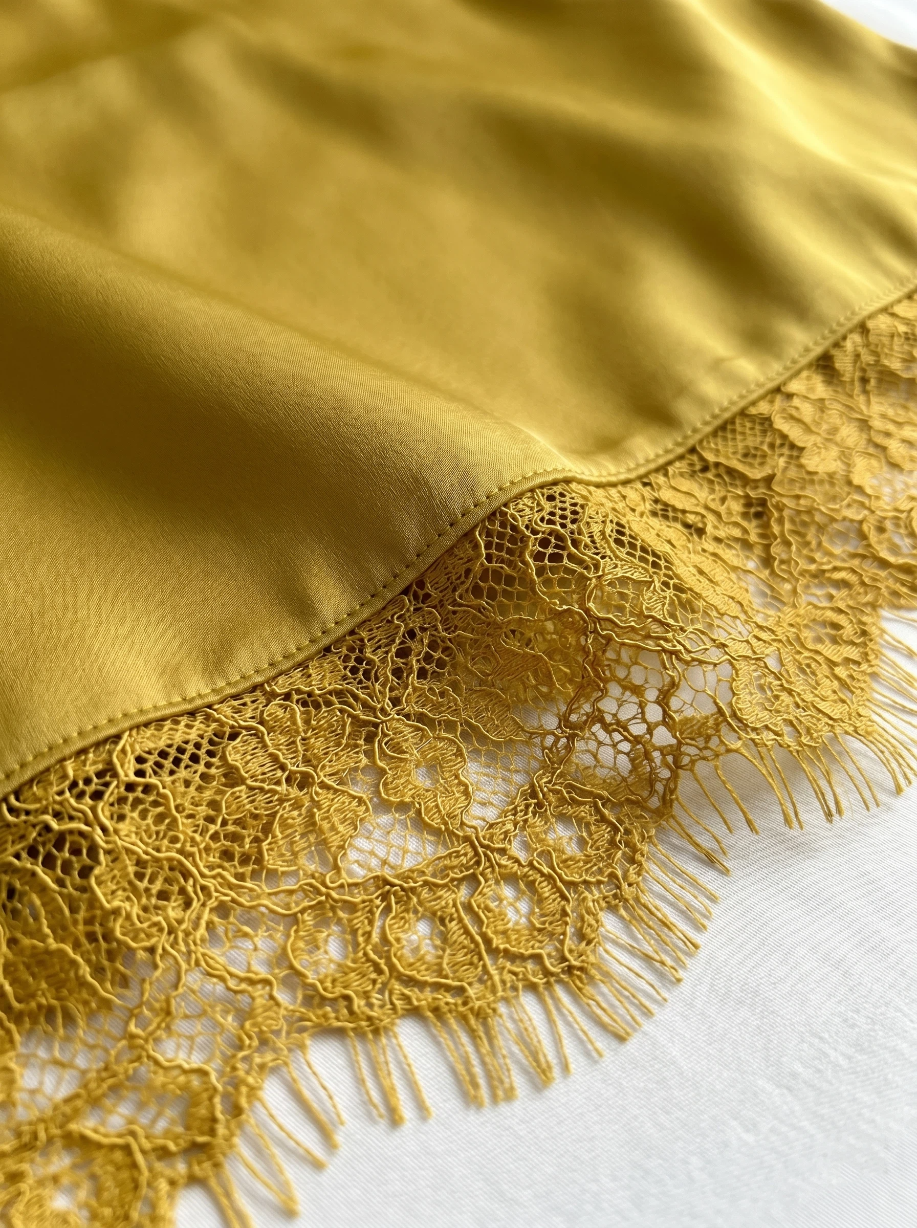 Close-up detail of luxurious gold satin fabric featuring a delicate, matching floral eyelash lace trim along the hem. A detailed shot highlighting smooth silk-like textures, elegant women's sleepwear edging, and luxury slip dress or lingerie materials.