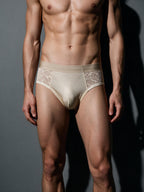 Male model wearing Sand Beige lace and silk men's brief, front view showcasing the smooth opaque contour pouch and sheer side panels against a dark backdrop.