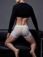 Back view of a male model leaning on a black sofa, wearing the ILLUSION white mesh lace trunks, showing the high-stretch, body-contouring silhouette that prevents rolling.
