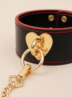 Close-up of the Velvet Fluxe luxury black leather cuff featuring a polished gold heart anchor, O-ring, and crimson red edge stitching. A premium intimate accessory for sophisticated bedroom aesthetics.
