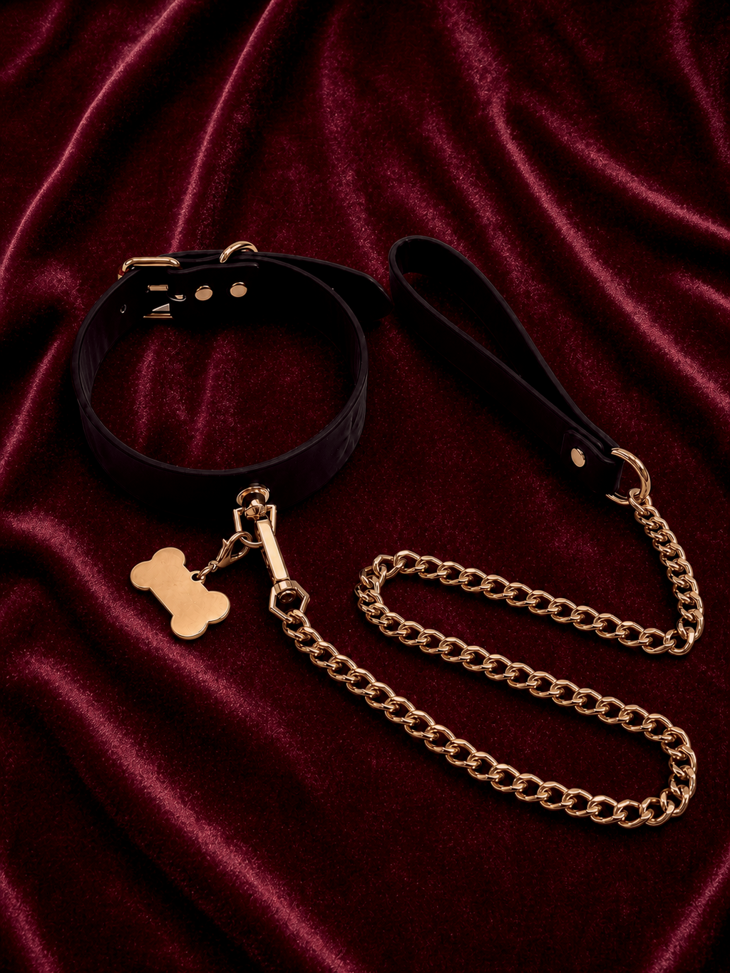 Flat lay of the Velvet Fluxe KIRA temperature-sensitive leather collar in its sleek midnight black state. Features a polished gold bone charm and heavy-duty gold link chain resting on deep red velvet. Luxury pet play intimate accessories.