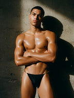Athletic male model wearing the Velvet Fluxe AURELIUS black thong with gold chain straps. Front view showcasing the masculine fit, glowing skin, and premium metallic hardware against a textured concrete wall.
