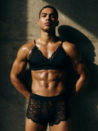 Muscular male model posing in the Velvet Fluxe NOCTURNE French black lace lingerie set. Front view featuring a gender-fluid black sheer lace triangle bralette and matching lace shorts with a delicate center bow.