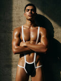 Athletic male model wearing the Velvet Fluxe black and white harness jockstrap. Front view highlighting the architectural white suspender straps and lifting black contoured pouch against a textured concrete wall.