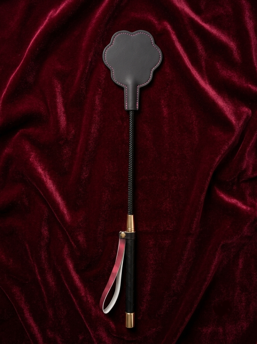 Velvet Fluxe FELINA luxury leather cat paw paddle. Back view featuring flat, smooth black leather with delicate pink contrast stitching and a weighted handle, resting on dark red velvet.